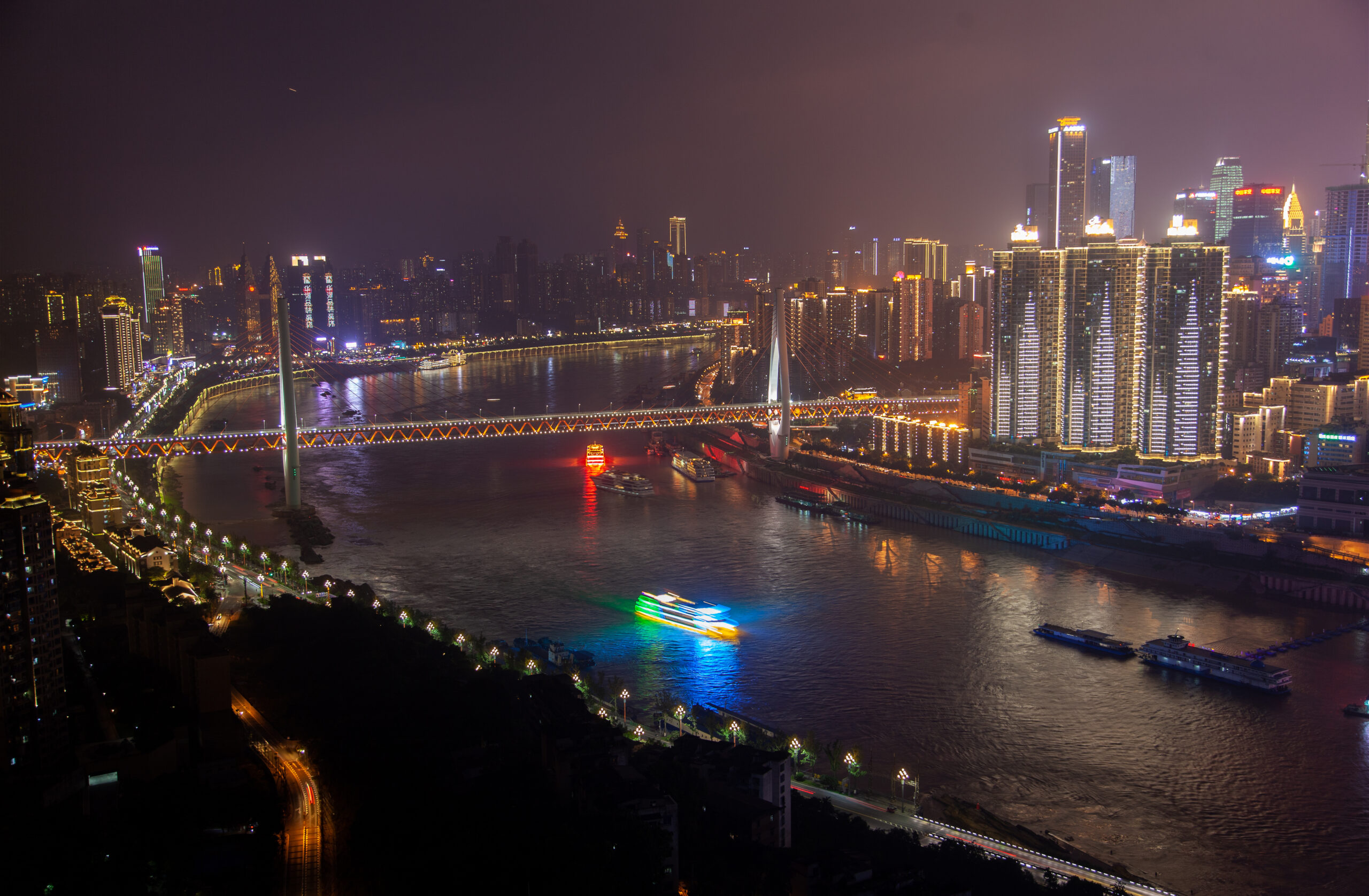 various motorboats sail along wide Yangtze river between illuminated Chongqing city districts buildings and skyscrapers in southwest China at night